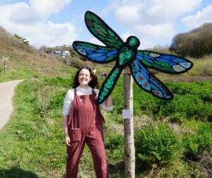 A young person stands in front of one of their dragonfly sculptures smiling at the camera in the sun.