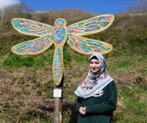 A young person stands in front of one of their dragonfly sculptures smiling at the camera in the sun.