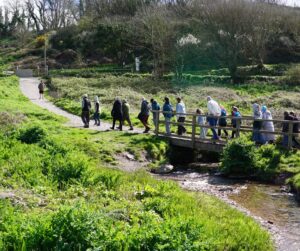 A large group of people walk across a bridge in the sun on the coast path.