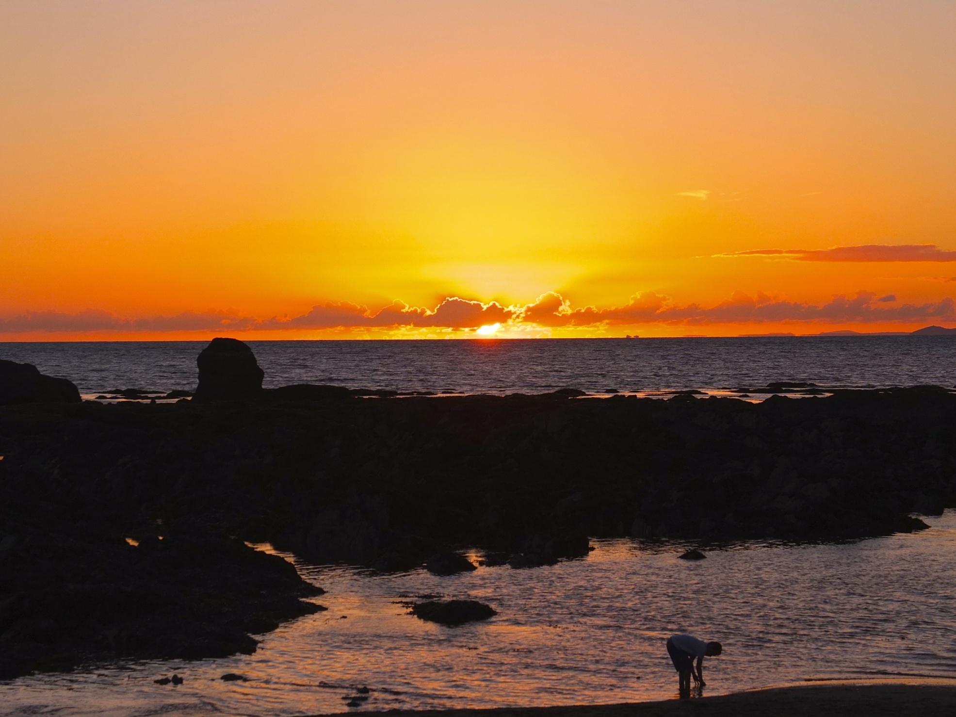 View from beach across the shallow water, the rocks and then out to the horizon where the sun is setting behind a few clouds