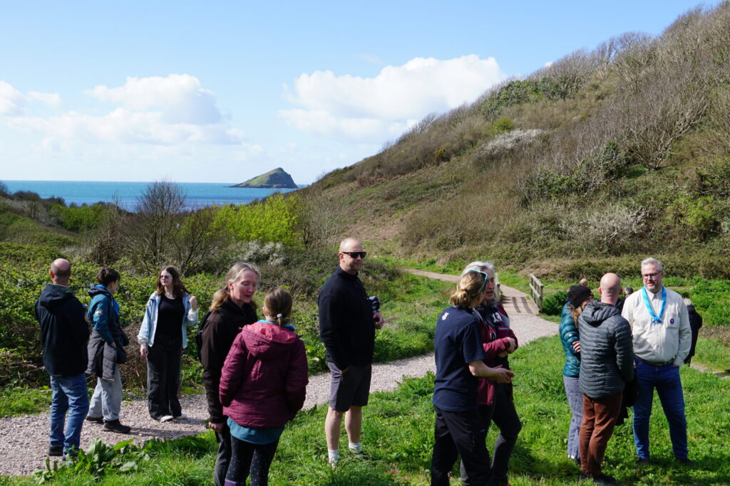 A group of people gather in a meadow with a valley and the sea behind them