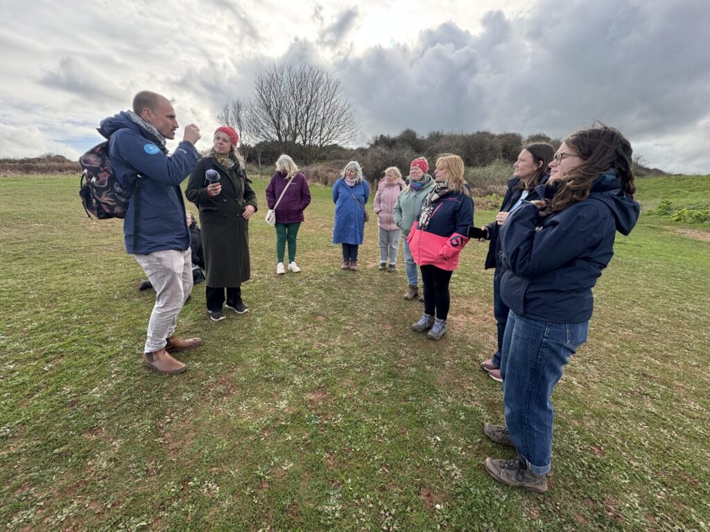A group of adults in a coastal clearing listening to someone speak about the area