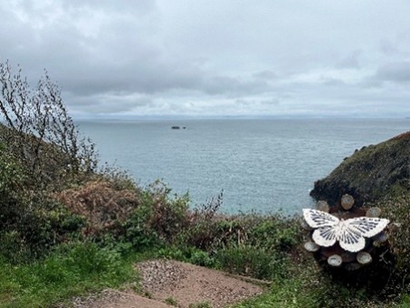 View from a cliff overlooking the sea with vegetation in the foreground . A large metalwork Silver-studded Blue sculpture is positioned to the right, just off the path