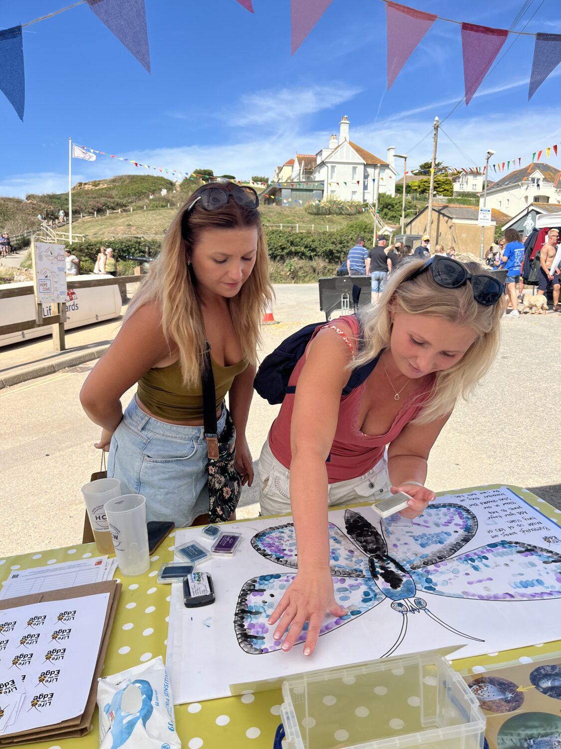 Two ladies colouring in a butterfly with inked fingers at an event in Hope Cove. In the background there is bunting, a few more people, a property atop grassy bank and blue skies.