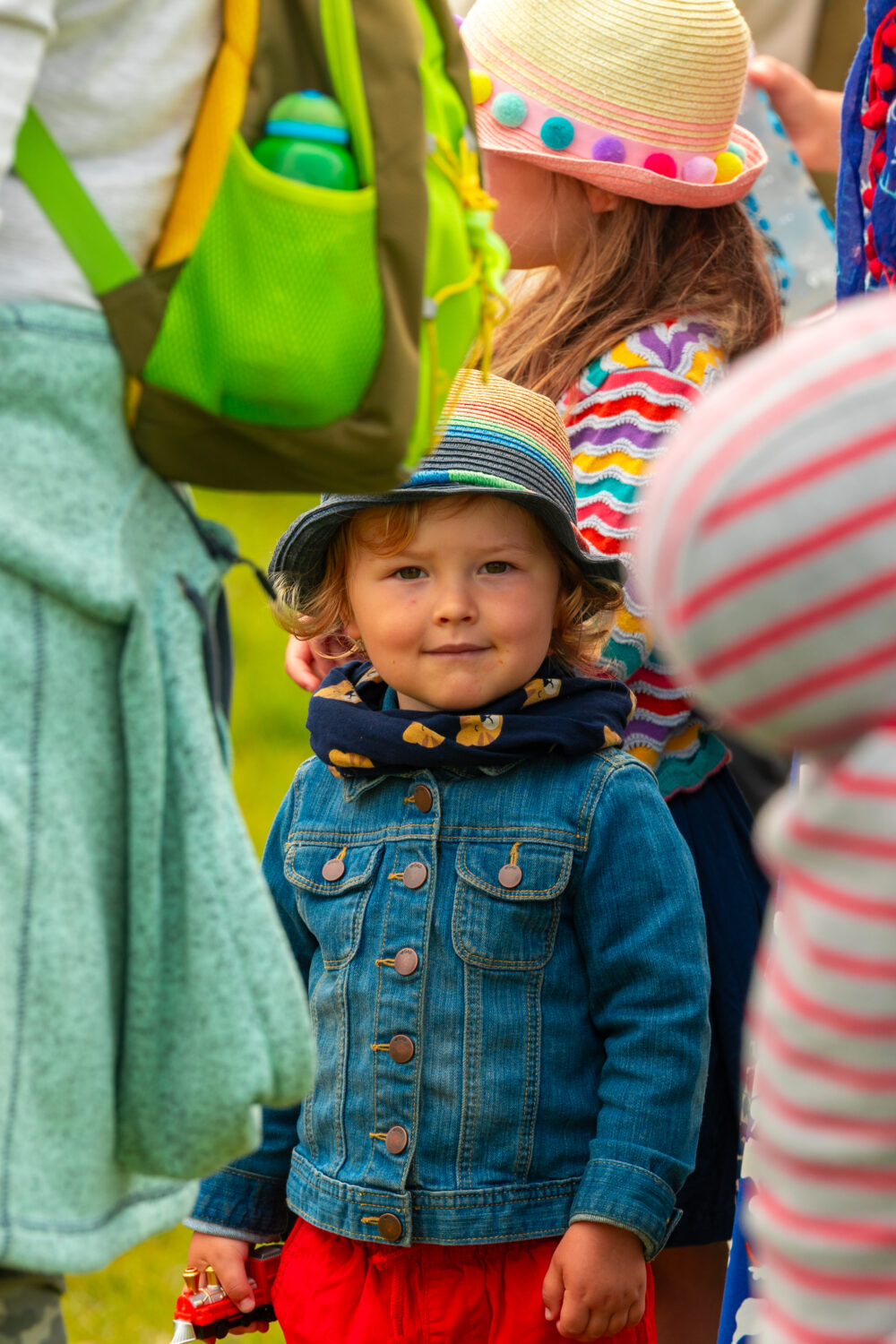 Close in shot of an adult and 3 children. One child is facing the camera, wearing a hat, denim jacket and red skirt holding a toy train